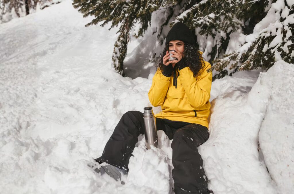 Woman in winter clothing enjoys a warm drink while sitting in snow-covered forest.